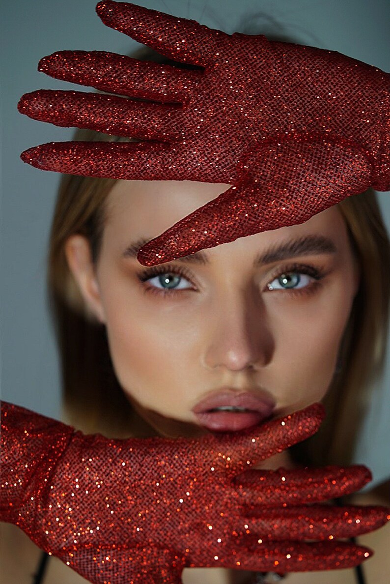 May include: A woman's face is partially obscured by a pair of red sparkly gloves. The gloves are held up to her face, covering her eyes and forehead. The gloves are made of a fine mesh fabric with a red glitter finish.