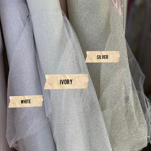 May include: Three rolls of sheer, glittery tulle fabric displayed side by side. The rolls are labeled with rectangular paper strips reading WHITE, IVORY, and SILVER. Each fabric features a fine, semi-transparent mesh texture with a sparkling finish.