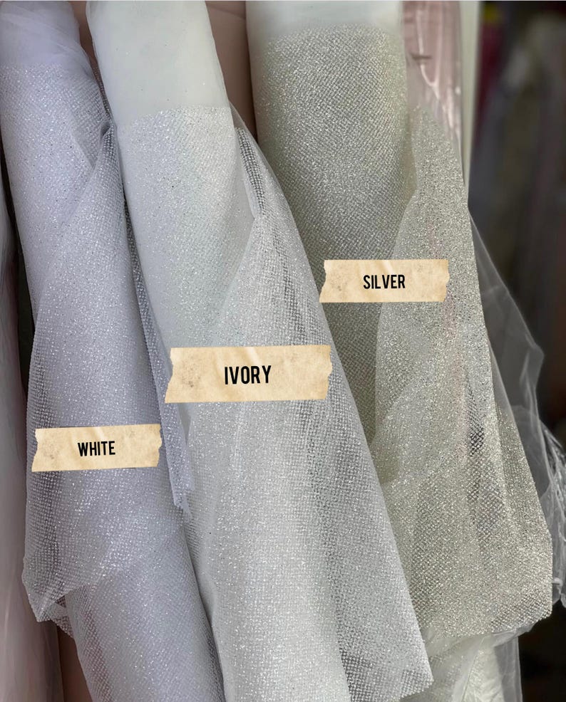 May include: Three rolls of sheer, glittery tulle fabric displayed side by side. The rolls are labeled with rectangular paper strips reading WHITE, IVORY, and SILVER. Each fabric features a fine, semi-transparent mesh texture with a sparkling finish.