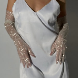 May include: A white satin slip dress featuring a V-neckline and thin spaghetti straps, paired with sheer, elbow-length opera gloves adorned with scattered silver sequins that catch the light, creating a shimmering and reflective appearance.
