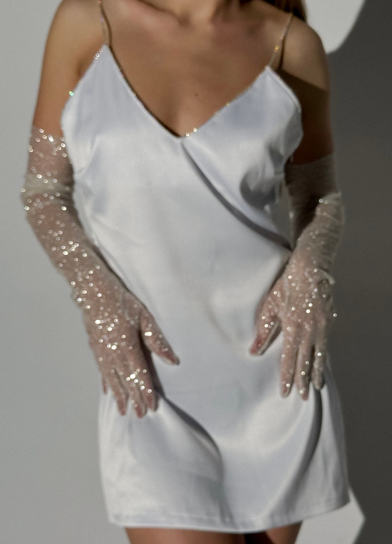 May include: A white satin slip dress featuring a V-neckline and thin spaghetti straps, paired with sheer, elbow-length opera gloves adorned with scattered silver sequins that catch the light, creating a shimmering and reflective appearance.