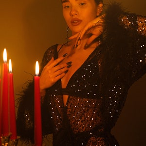 May include: A woman wearing a black sequined robe with feather trim, standing in front of lit candles. The robe is sheer and has a plunging neckline.