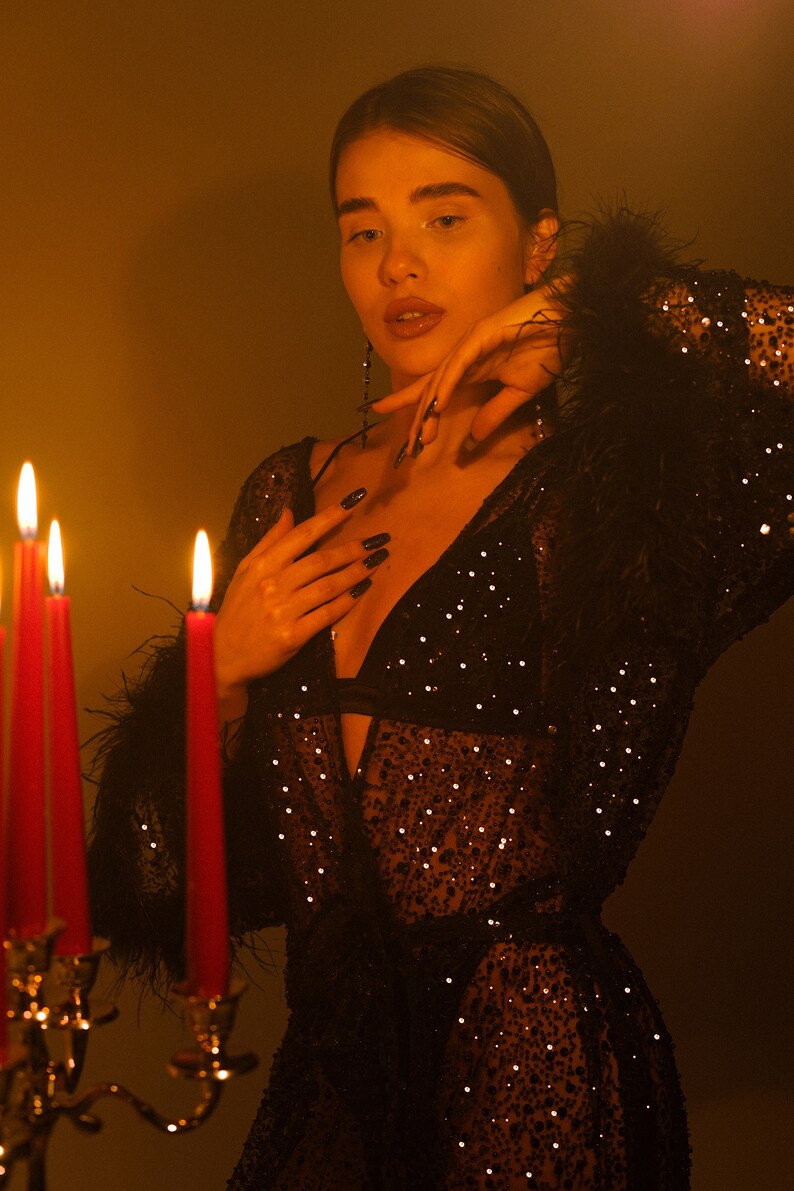May include: A woman wearing a black sequined robe with feather trim, standing in front of lit candles. The robe is sheer and has a plunging neckline.