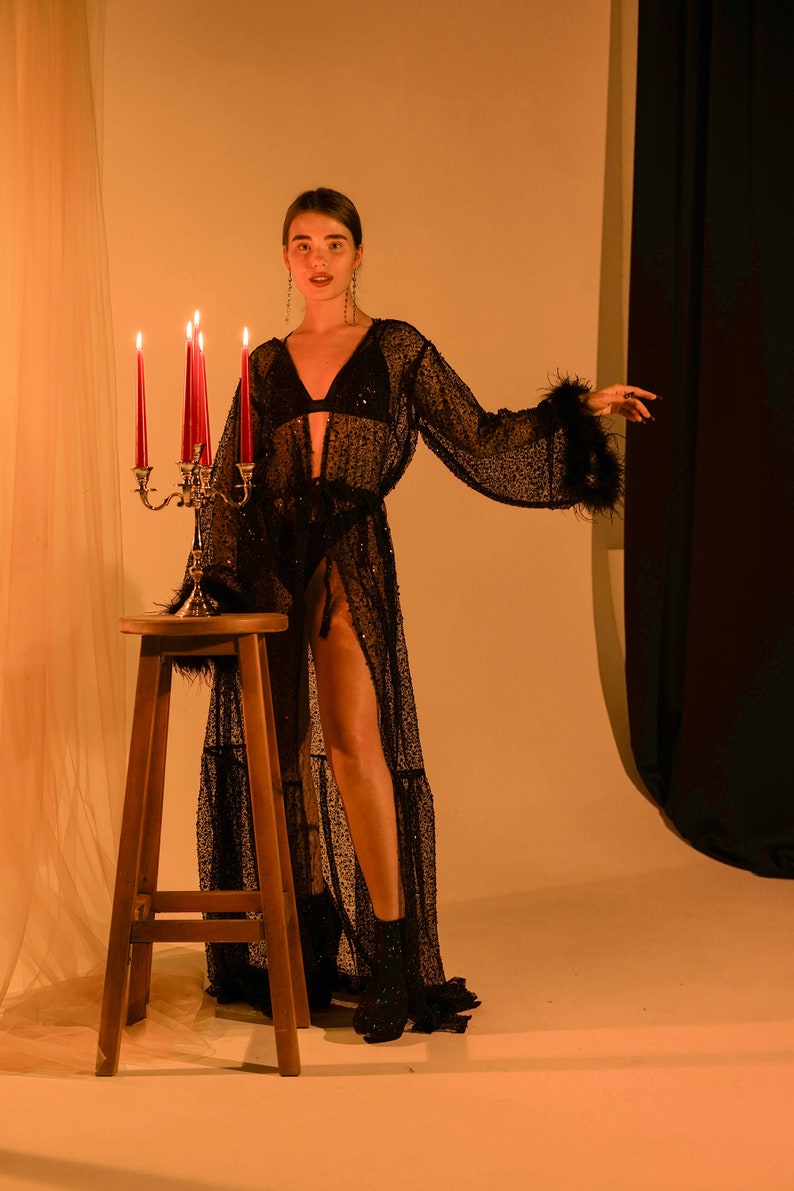 May include: A woman wearing a black sequined robe with feather trim, standing in front of a candle holder with lit candles. The background is a light orange color.