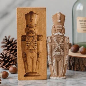 May include: A wooden mould and a decorative soldier figurine. The mould is rectangular with a carved soldier design. The figurine is tan with a detailed soldier design. The image also includes pine cones, nuts, and a glass bottle.
