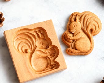 May include: A wooden squirrel-shaped cookie mold and a baked cookie. The mold is rectangular with a carved squirrel design. The cookie is light brown, matching the color of the mold. Acorns and pinecones are scattered around the mold and cookie.