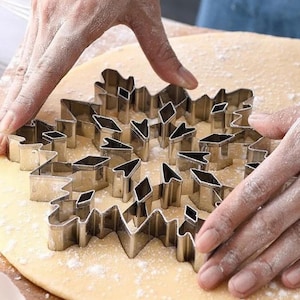 May include: A snowflake-shaped cookie cutter made of silver metal is pressed into a circle of dough dusted with flour. The image shows the process of making cookies, with hands pressing the cutter into the dough.