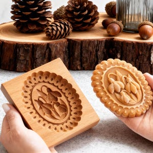 Può includere: Uno stampo per biscotti in legno e un biscotto cotto con un disegno floreale sono tenuti in mano. Lo stampo è rettangolare con un motivo floreale ovale. Il biscotto ha un design simile. Pigne, ghiande e una bottiglia decorativa sono su una fetta di legno.