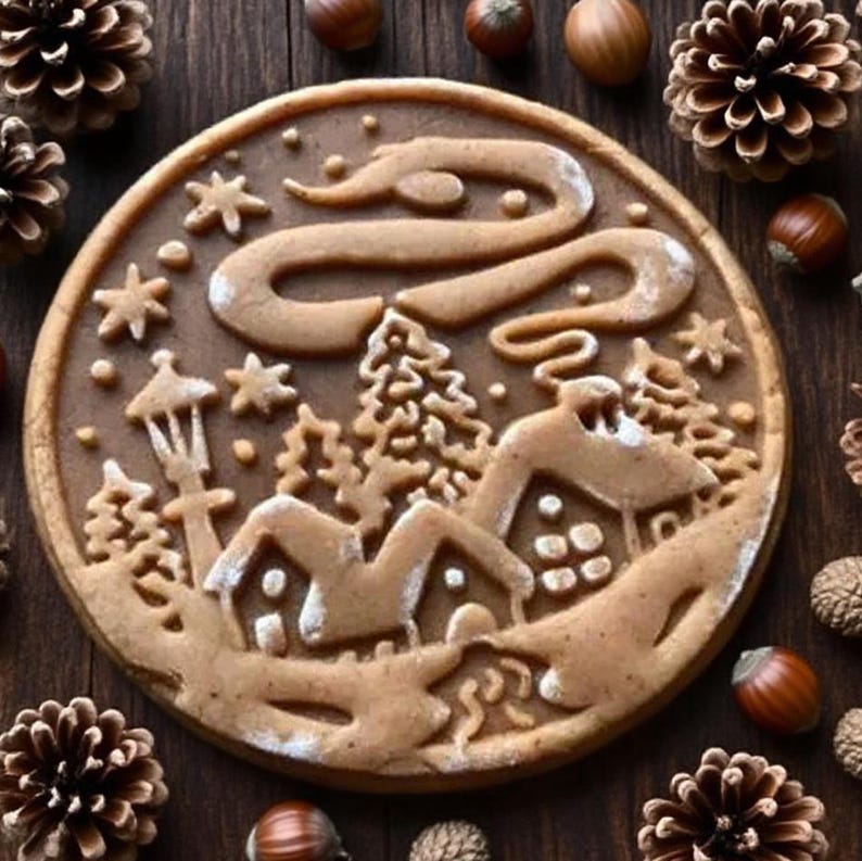May include: A round gingerbread cookie with a winter scene design. The cookie features a detailed depiction of houses, trees, stars, and a winding path, all dusted with white accents. Pine cones and hazelnuts surround the cookie.