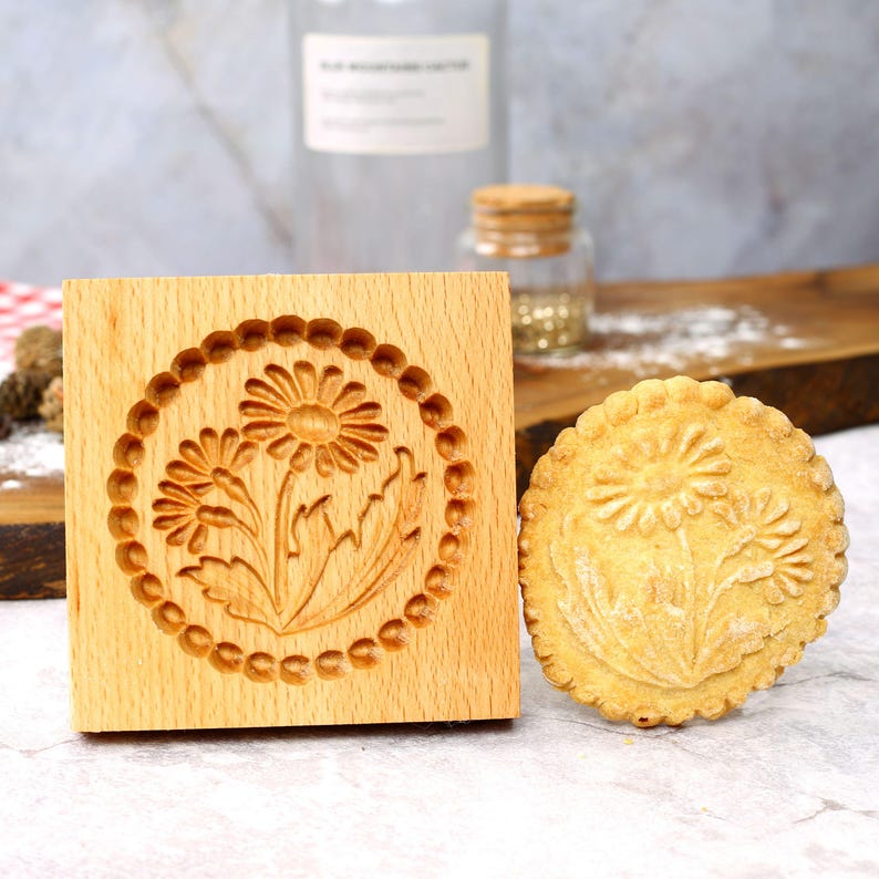 May include: A wooden cookie cutter with a daisy design. The cutter is next to a cookie with the same daisy design.
