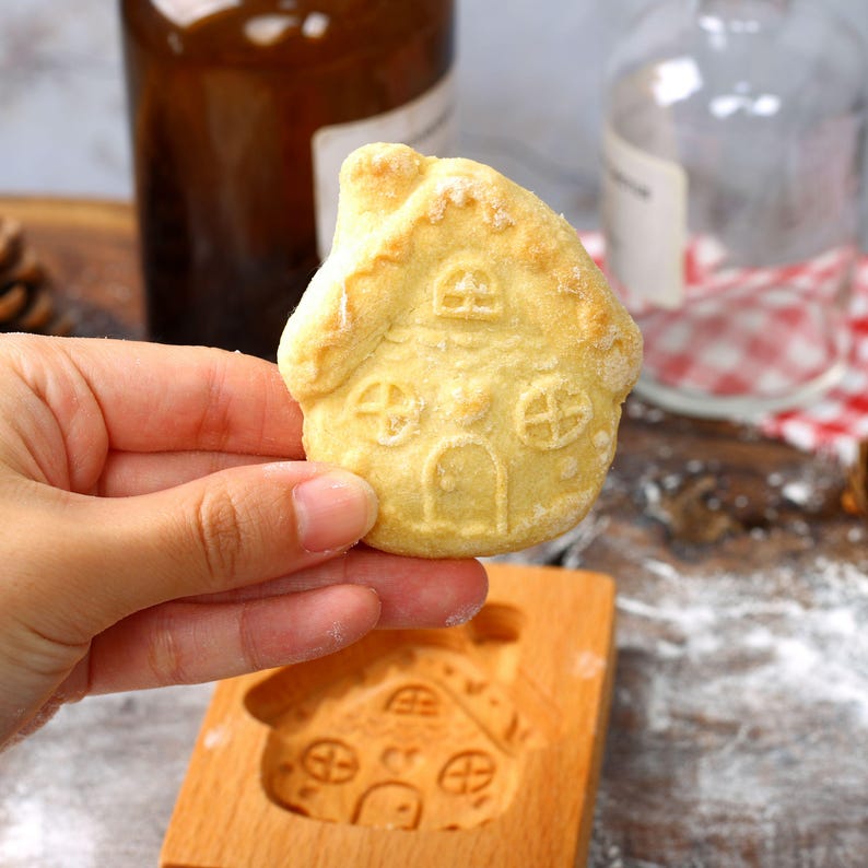 Op de afbeelding: Een lichtbruine gingerbreadkoek in de vorm van een huis met een bevroren dak en ramen. De koek wordt in een hand boven een houten koekjesvormpje gehouden.