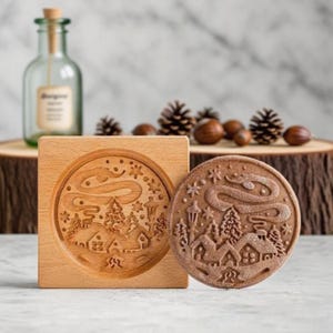 May include: A wooden cookie mold and a pressed cookie with a winter village design. The mold is square with a circular indentation. The cookie is round and brown, with a detailed relief of houses, trees, and a swirling sky.
