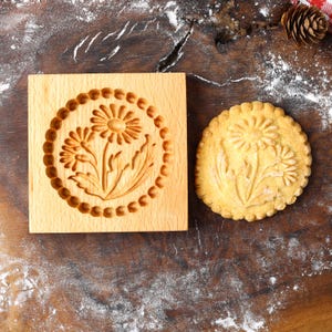 May include: A wooden cookie cutter with a floral design, and a cookie with the same design. The cookie cutter is square and has a raised design of two flowers with leaves. The cookie is light brown and has a similar design.