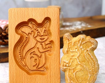 May include: A wooden cookie cutter in the shape of a squirrel. The cutter is made of light brown wood and has a detailed design of a squirrel with a bushy tail. Next to the cutter is a cookie that has been cut out using the cutter. The cookie is a light brown color and has a similar design to the cutter.