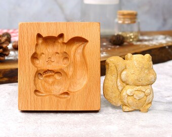 May include: A wooden mold for making cookies or other treats in the shape of a squirrel. The mold is made of light brown wood and has a detailed design of a squirrel holding a nut. A cookie made with the mold is shown next to it.
