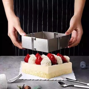 Puede incluir: Un molde cuadrado para pasteles de acero inoxidable se sostiene sobre un pastel rectangular decorado con fresas y crema. El pastel se encuentra sobre un mantel individual de papel blanco, con un tenedor, un cuchillo y una cuchara cerca. El fondo es un delantal a rayas blanco y negro.