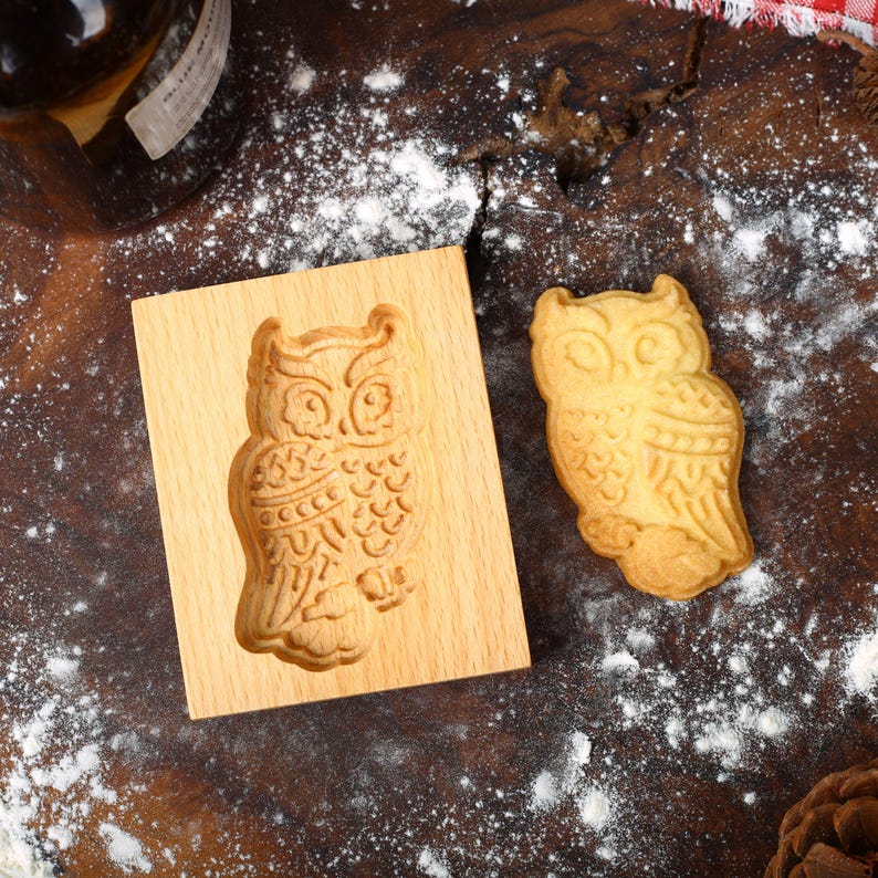 May include: A wooden cookie cutter shaped like an owl, with a cookie in the shape of an owl next to it. The cookie cutter and cookie are on a brown wooden surface covered in white flour.