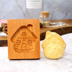 Op de afbeelding: Een houten gingerbread house koekjesvormpje met een gingerbread house koekje ernaast. De koekjesvormpje is gemaakt van lichtbruin hout en heeft een gedetailleerd ontwerp van een gingerbread house met een schoorsteen, ramen en een deur. De koekje is licht goudkleurig bruin en heeft een vergelijkbaar ontwerp als de koekjesvormpje.
