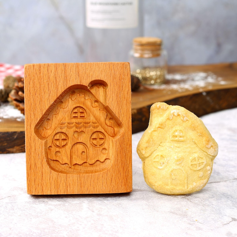 Op de afbeelding: Een houten gingerbread house koekjesvormpje met een gingerbread house koekje ernaast. De koekjesvormpje is gemaakt van lichtbruin hout en heeft een gedetailleerd ontwerp van een gingerbread house met een schoorsteen, ramen en een deur. De koekje is licht goudkleurig bruin en heeft een vergelijkbaar ontwerp als de koekjesvormpje.