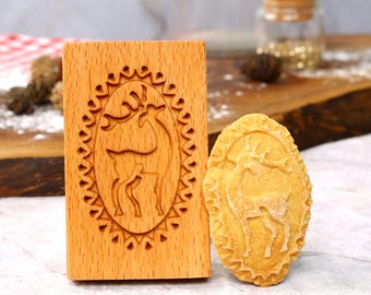 May include: A wooden cookie stamp with a reindeer design and a cookie with the same design. The cookie stamp is made of light brown wood and has a rectangular shape. The cookie is a light brown color and has an oval shape.