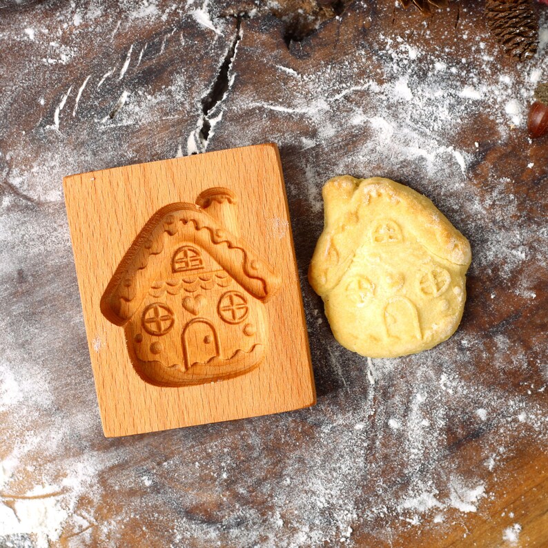 Op de afbeelding: Een houten gingerbread house koekjesvormpje met een gingerbread house koekje ernaast op een bruin houten oppervlak bedekt met witte bloem.