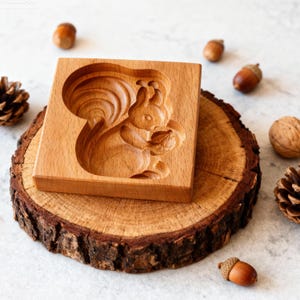 May include: A square, wooden mold with a carved squirrel design holding a nut. The mold is light brown and sits on a wooden slice, surrounded by acorns, a walnut, and a pinecone. The squirrel design is detailed and the wood grain is visible.