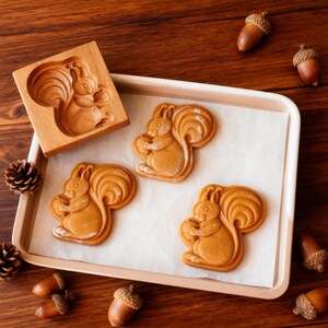 May include: A wooden squirrel-shaped cookie mold next to three squirrel-shaped cookies on a baking sheet. The cookies are light brown, and the mold is made of light-colored wood. Acorns are scattered around the baking sheet.