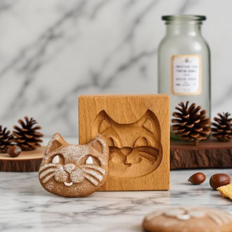 May include: A wooden cat-shaped cookie mold and a baked cookie. The mold is square and features a carved cat face. The cookie is light brown with white details. A glass bottle and pinecones are in the background.