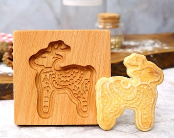 May include: A wooden cookie mold with a carved reindeer design next to a baked cookie in the same shape. The mold is made of light-colored wood and features detailed carving. The cookie is golden brown and has a detailed pattern.