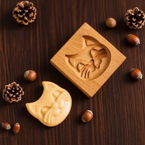 May include: A wooden cat-shaped cookie mold with a matching cookie. The mold is square with a carved cat face design. The cookie is light brown and has the same cat face design. Pinecones and hazelnuts are scattered around.