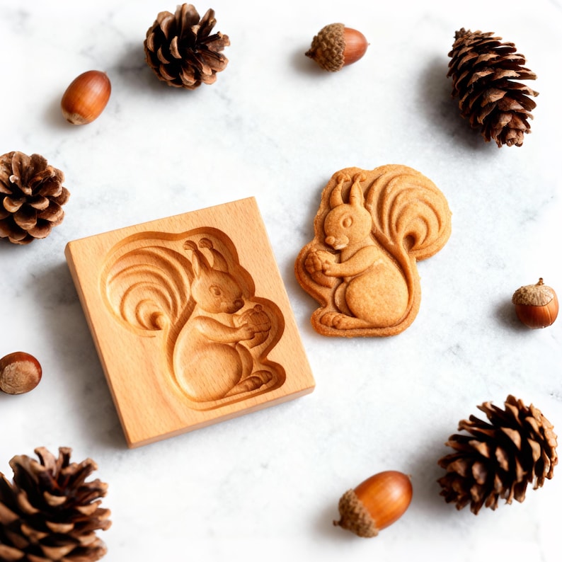 May include: A wooden squirrel-shaped cookie mold and a baked cookie. The mold is rectangular with a carved squirrel design. The cookie is light brown, matching the color of the mold. Acorns and pinecones are scattered around the mold and cookie.