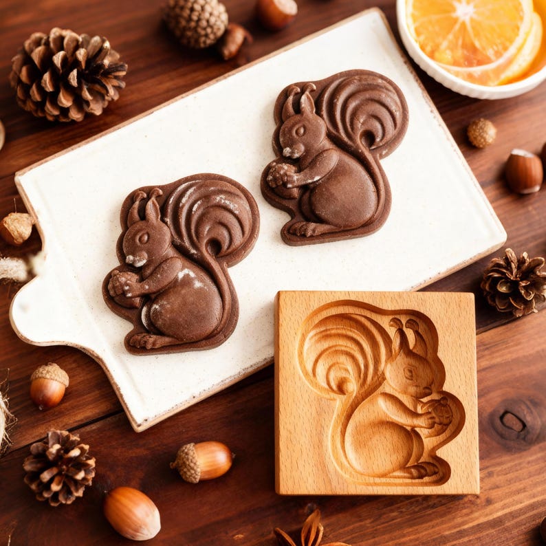 May include: Three chocolate squirrel-shaped cookies on a white rectangular plate, alongside a wooden squirrel mold. The cookies are dark brown, and the mold is light brown. Acorns and pinecones are scattered around the plate on a wooden surface.