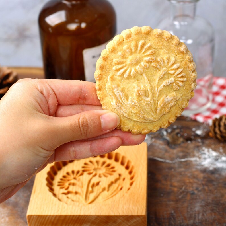 May include: A close-up of a light brown cookie with a floral design pressed into the surface. The cookie is being held by a hand and is in front of a wooden cookie mold with the same floral design.