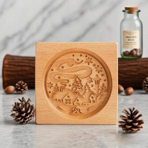 May include: A square, light-colored wooden block with a circular, engraved winter scene. The scene includes houses, trees, and a swirling design, possibly representing the Northern Lights. A glass jar with a cork top and pine cones are also visible.