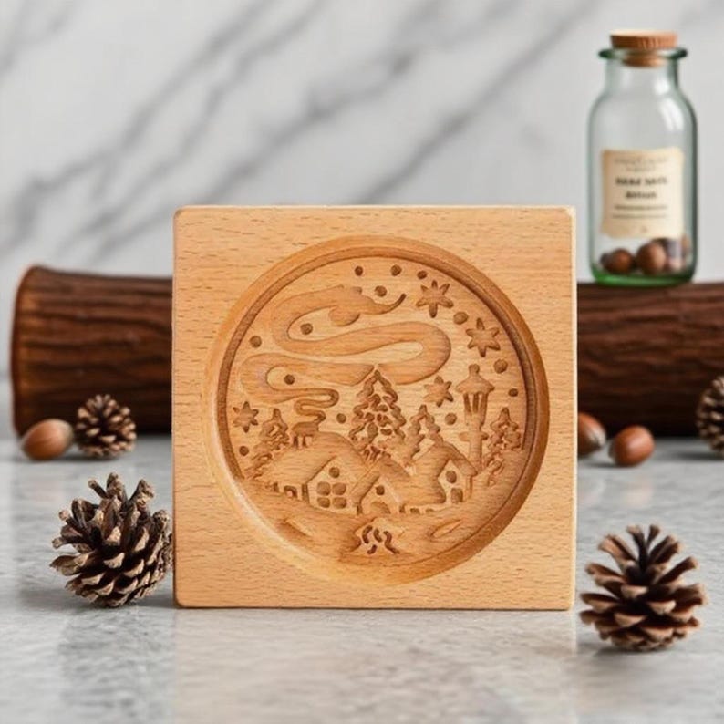 May include: A square, light-colored wooden block with a circular, engraved winter scene. The scene includes houses, trees, and a swirling design, possibly representing the Northern Lights. A glass jar with a cork top and pine cones are also visible.