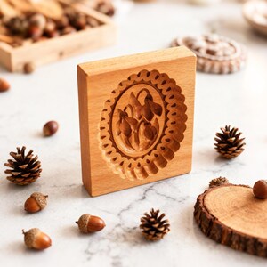 K&ouml;nnte beinhalten: Eine Holzkeksform mit einem floralen Design, das in ihre Oberfl&auml;che geschnitzt ist. Die Form ist rechteckig mit einem gewellten Rand um das florale Muster. Eicheln, Tannenzapfen und ein Holztablett mit N&uuml;ssen sind um die Form verstreut.
