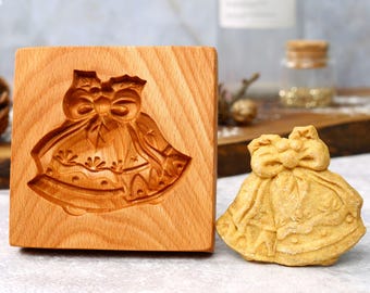 Könnte beinhalten: Holzförmchen in Form einer Glocke mit Schleife. Der Ausstecher ist aus hellem braunem Holz und hat ein detailliertes Design. Ein mit dem Ausstecher gemachter Keks ist daneben abgebildet.