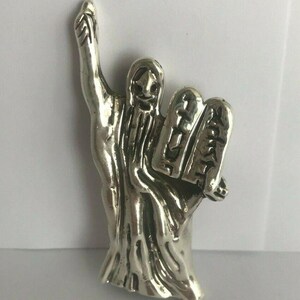 May include: A silver-colored metal figurine of a person holding two tablets with writing on them. The person is wearing a long robe and has a beard. The figurine is standing on a white surface.