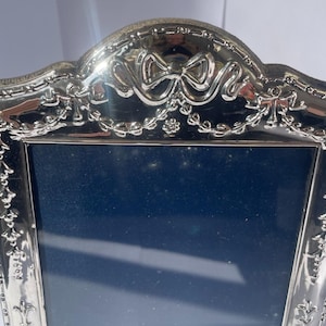 May include: A silver-toned picture frame with a polished metallic finish. The frame features an arched top adorned with a raised bow motif and intricate garland detailing along the border. The central display area is a dark, solid navy blue colour.