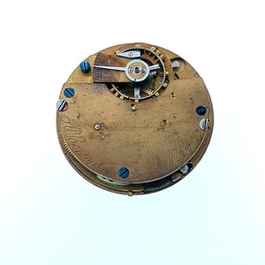 May include: Close-up of a vintage watch movement. The brass mechanism features intricate gears, screws, and a balance wheel. The text "Best London Make" and "J. Wilson" are visible. The watch has a circular shape and is set against a white background.