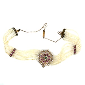 May include: A vintage pearl choker necklace with multiple strands. The necklace features a central floral pendant with red and green gemstones, and rectangular accents with red gemstones. The clasp and chain are gold-toned.
