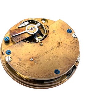 May include: Close-up of a vintage watch movement with intricate gears and components. The brass-colored mechanism features the text "West London Wake" and "J.W. Benson". Blue screws and other small details are visible.