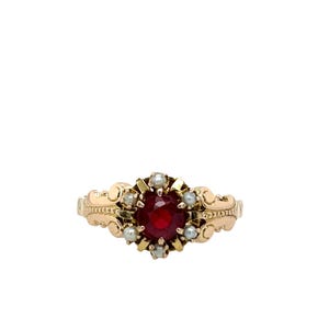 Puede incluir: Un anillo de oro con una gema roja oscura, redonda y grande en el centro, rodeada de perlas más pequeñas. El anillo tiene detalles dorados ornamentados y ondulados en la banda.