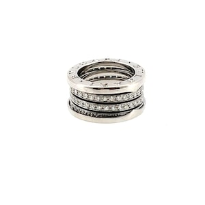 May include: A silver-toned metal ring featuring a multi-band design with a central row of small, clear, sparkling stones. The top and bottom edges of the ring are smooth and engraved with the text BVLGARI on both the top and bottom rims.
