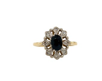 May include: A vintage gold ring featuring a dark blue oval gemstone at the center, surrounded by a halo of smaller, clear gemstones. The ring has a detailed, floral-inspired design around the central stone.