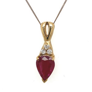 Könnte beinhalten: Eine goldene Halskette mit Anhänger, die einen birnenförmigen, tiefroten Rubin-Edelstein aufweist. Der Anhänger ist mit drei kleinen, runden Diamanten verziert und hängt an einer feinen Goldkette. Der Schmuck ist vor einem weißen Hintergrund platziert.