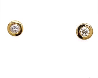 Puede incluir: Un par de pendientes de oro con tachuelas, cada uno con un solo diamante redondo y transparente engastado en un bisel. Los pendientes se muestran sobre un fondo blanco, destacando la simple elegancia de la joyería.