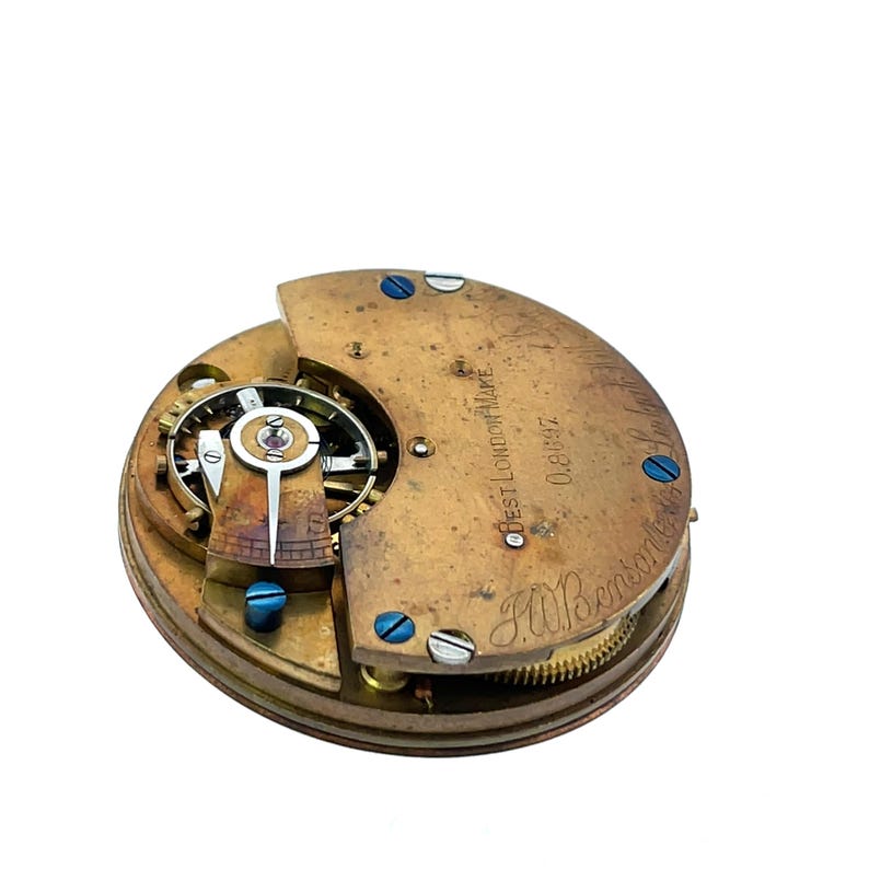 May include: Close-up of a vintage watch movement with exposed gears and intricate mechanisms. The brass-colored components are secured with blue screws. Text on the watch reads "BEST LONDON MAKE" and "F.W.Benson".
