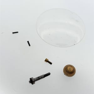 May include: A collection of watch parts, including small screws, a gear, and a decorative gold-colored knob. A clear, circular magnifying glass is also present. The parts are scattered on a white surface.