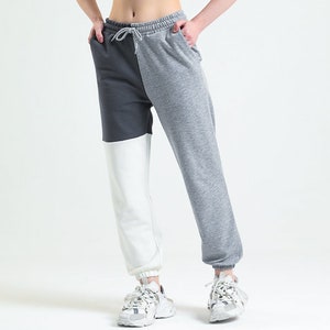 Può includere: Una donna indossa un top corto bianco e pantaloni della tuta a blocchi di colore. I pantaloni della tuta sono divisi in tre sezioni verticali: grigio scuro, bianco e grigio chiaro. Indossa anche scarpe da ginnastica bianche.